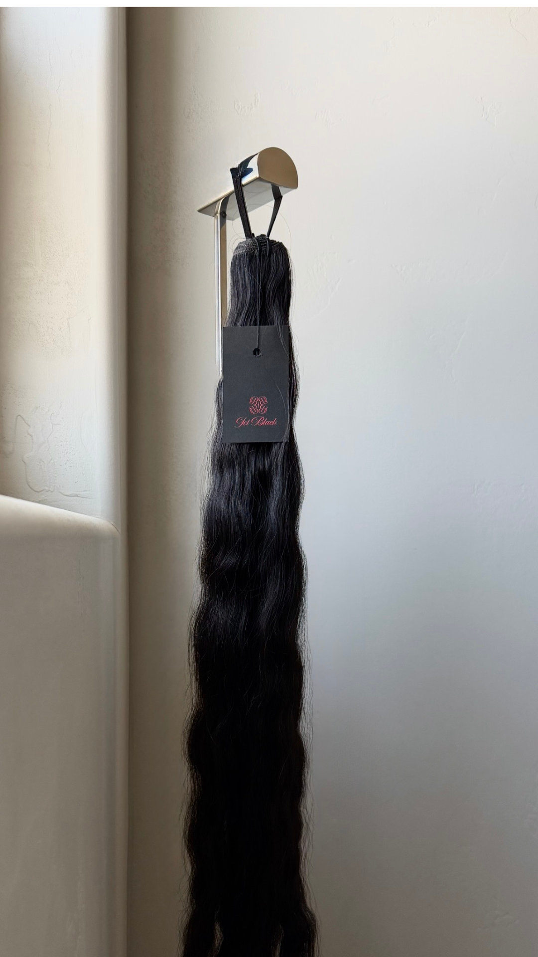 Hair extensions hanging on a product display with a Jet Black product tag against a white wall