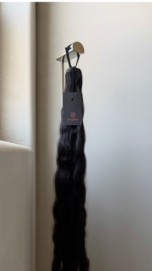 Hair extensions hanging on a product display with a Jet Black product tag against a white wall